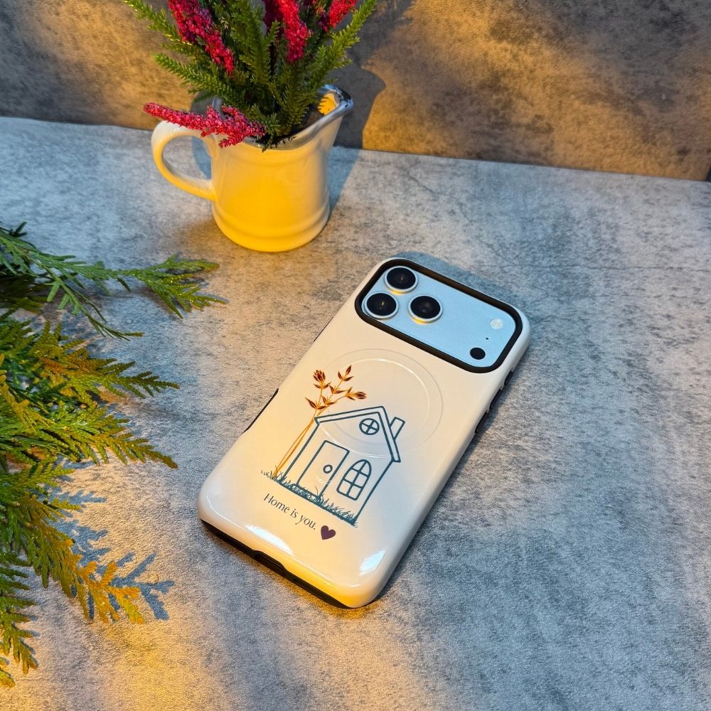 White iPhone case featuring a minimalist house illustration and 'Home is you' text, displayed on a grey stone surface next to a white pitcher of pink flowers.